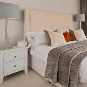 Bedroom with white bedside table, decorative cushions and throws, big statement lamps and beige carpet