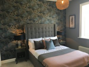 Grey fabric bed with green, grey and pink cushions and throws, art prints, bold patterned wallpaper and statement lighting