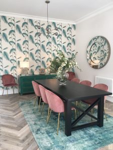 Black wooden dining table with velvet pink chairs, green sideboard, patterned wallpaper and statement mirror