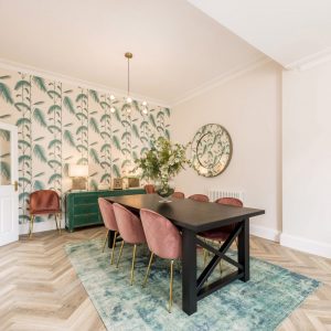 Black wooden dining table with velvet pink chairs, green sideboard, patterned wallpaper and statement mirror