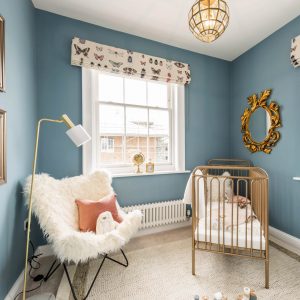 Luxury baby's nursery with gold metal crib, fluffy white armchair, blue walls, gold statement mirror, lamp and framed photos